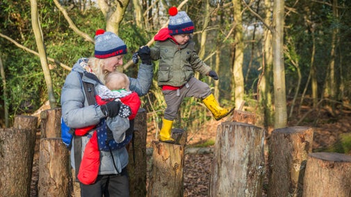 Woman with a baby in a sling, holding the hand of a toddler as he walks across logs on the advenure area